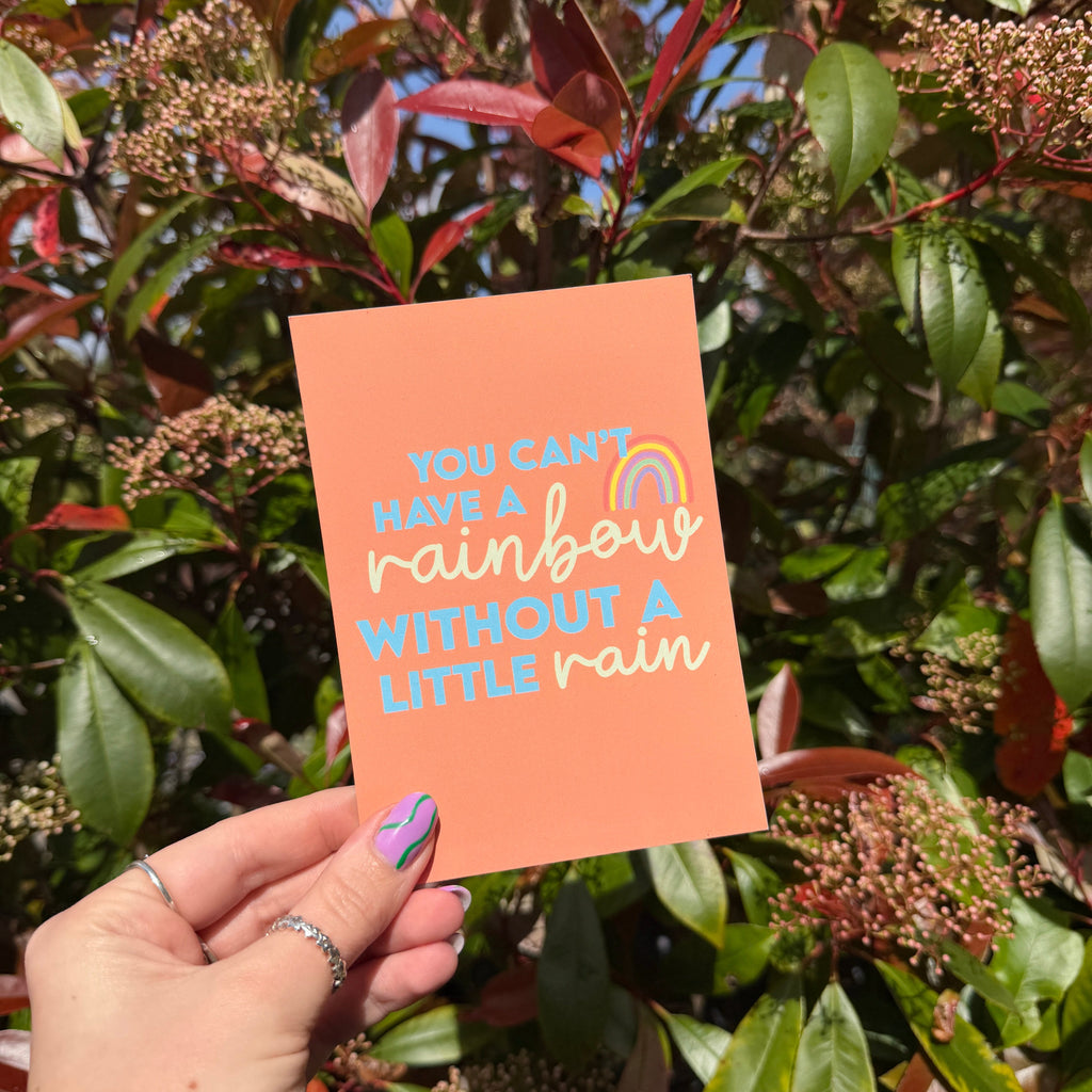 Hand holding an orange postcard with words 'You can't have a rainbow without a little rain' on a leafy background