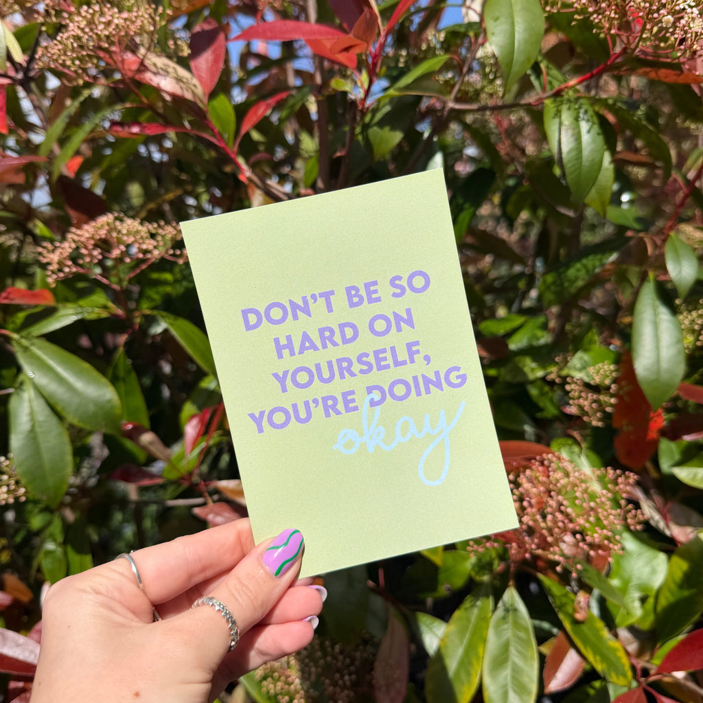 Hand holding a green postcard with the words 'Don't be so Hard on Yourself, You're Doing Okay' against a leafy background