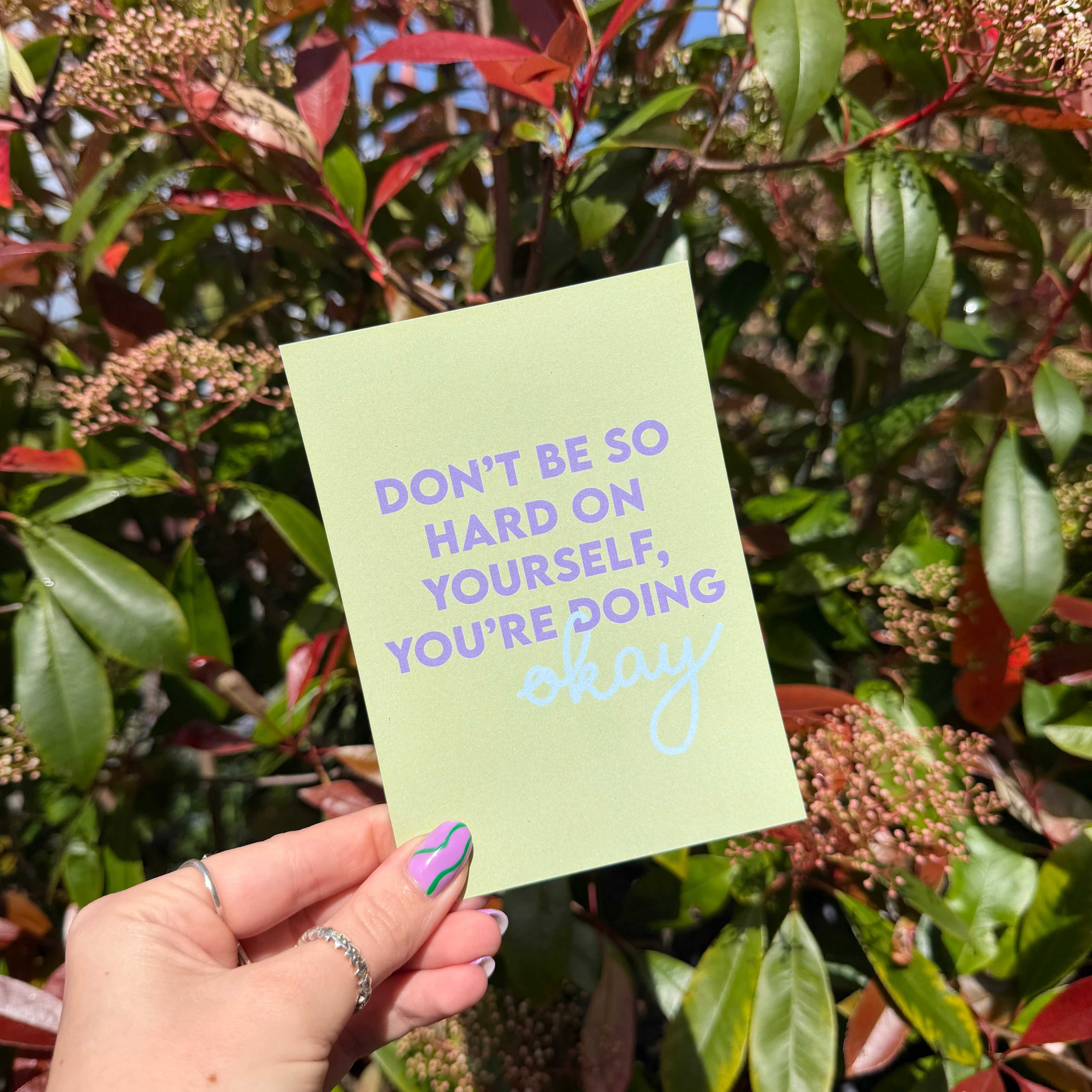 Hand holding a green postcard with the words 'Don't be so Hard on Yourself, You're Doing Okay' against a leafy background