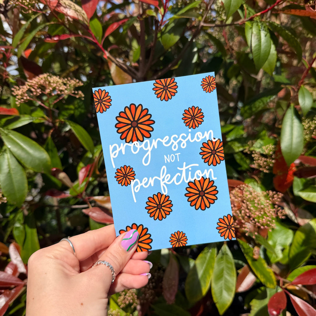 Hand holding a postcard with a floral design and the words 'Progression not perfection' against a leafy background