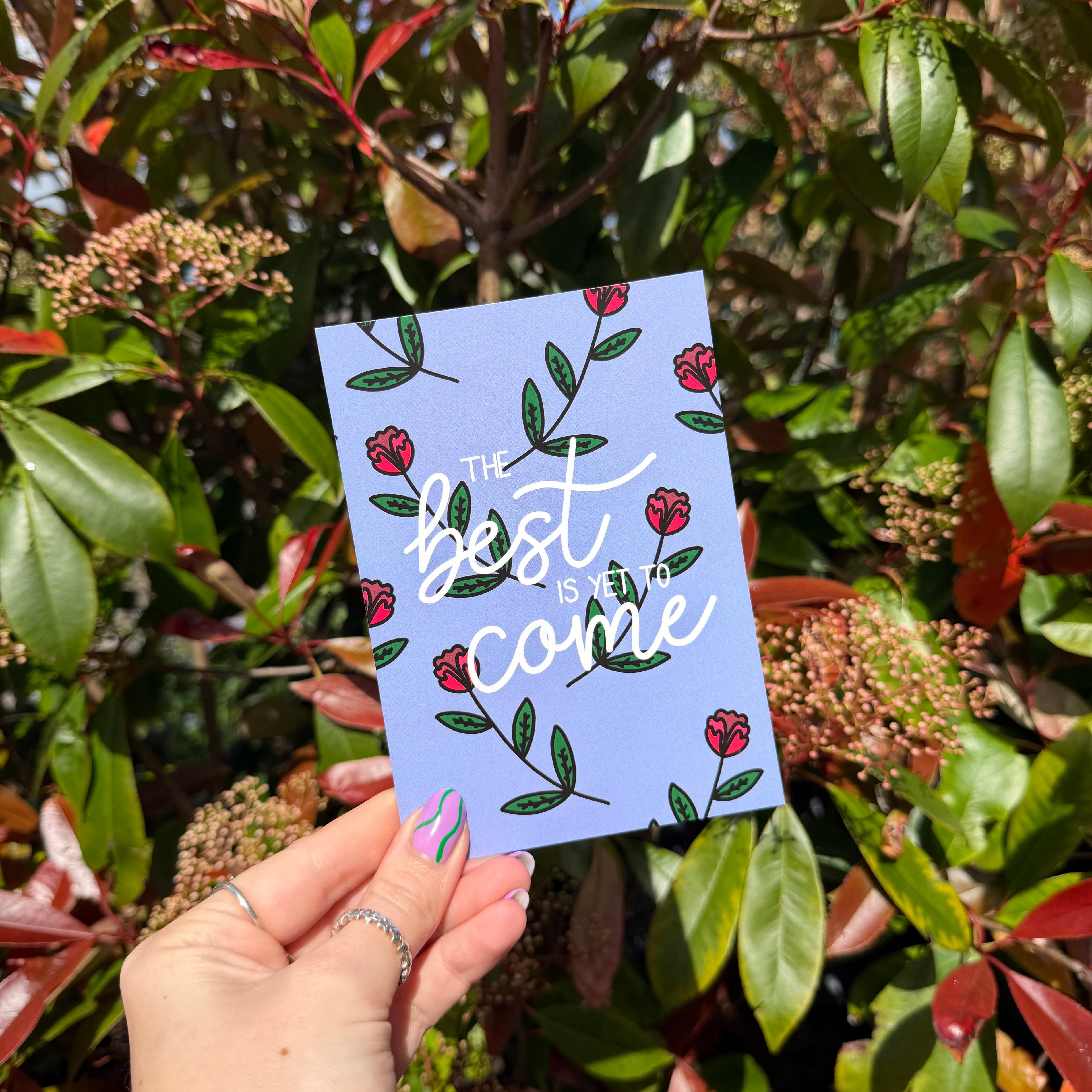 Hand holding a purple post card with floral design and words 'The best is yet to come' amidst green foliage