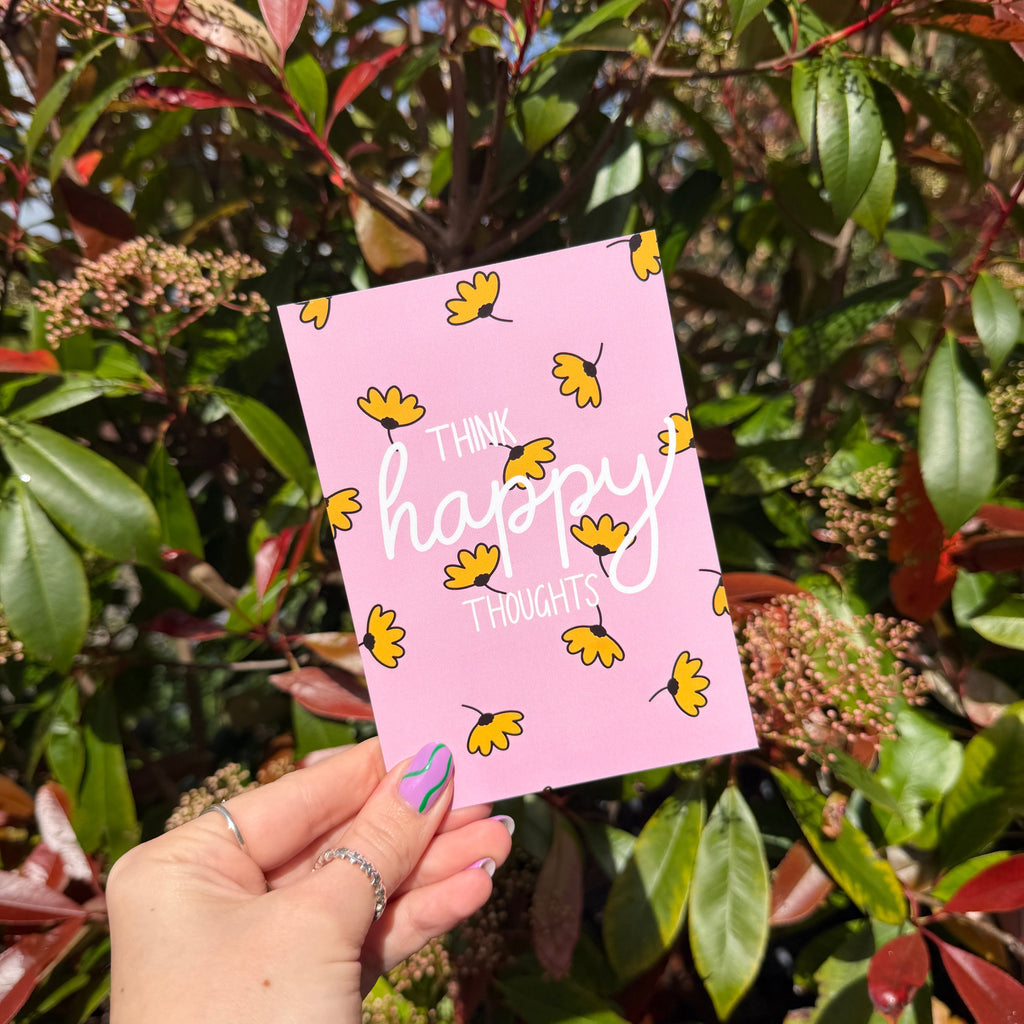 Hand holding a pink card with yellow flowers and the words 'Think Happy Thoughts' in front of green foliage