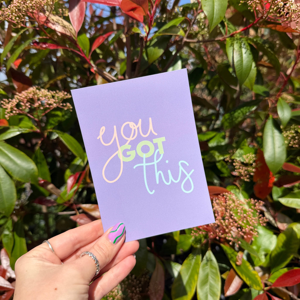 Hand holding a purple postcard with 'you got this' message among green foliage