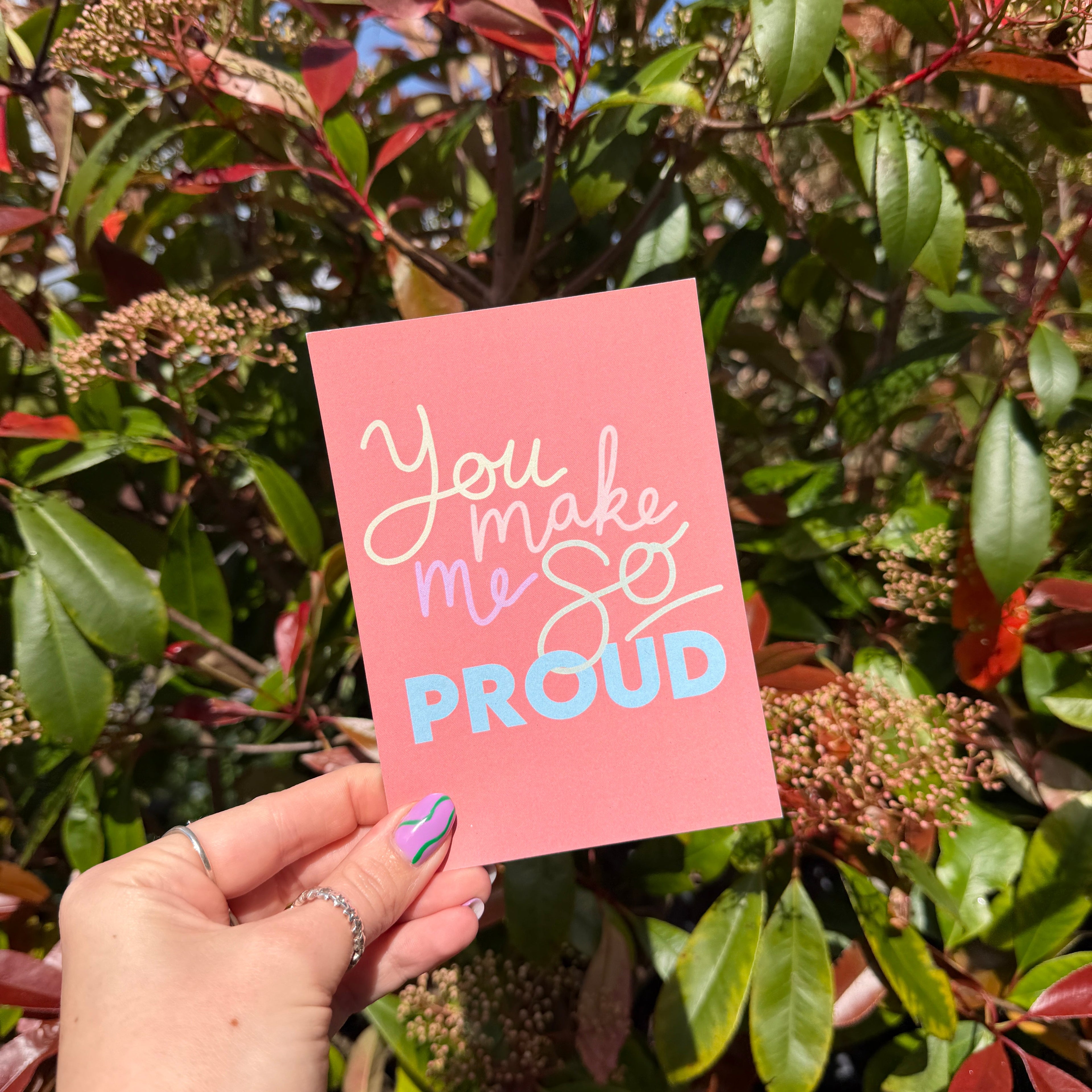 Hand holding a coral postcard with 'You make me proud' text against a leafy background
